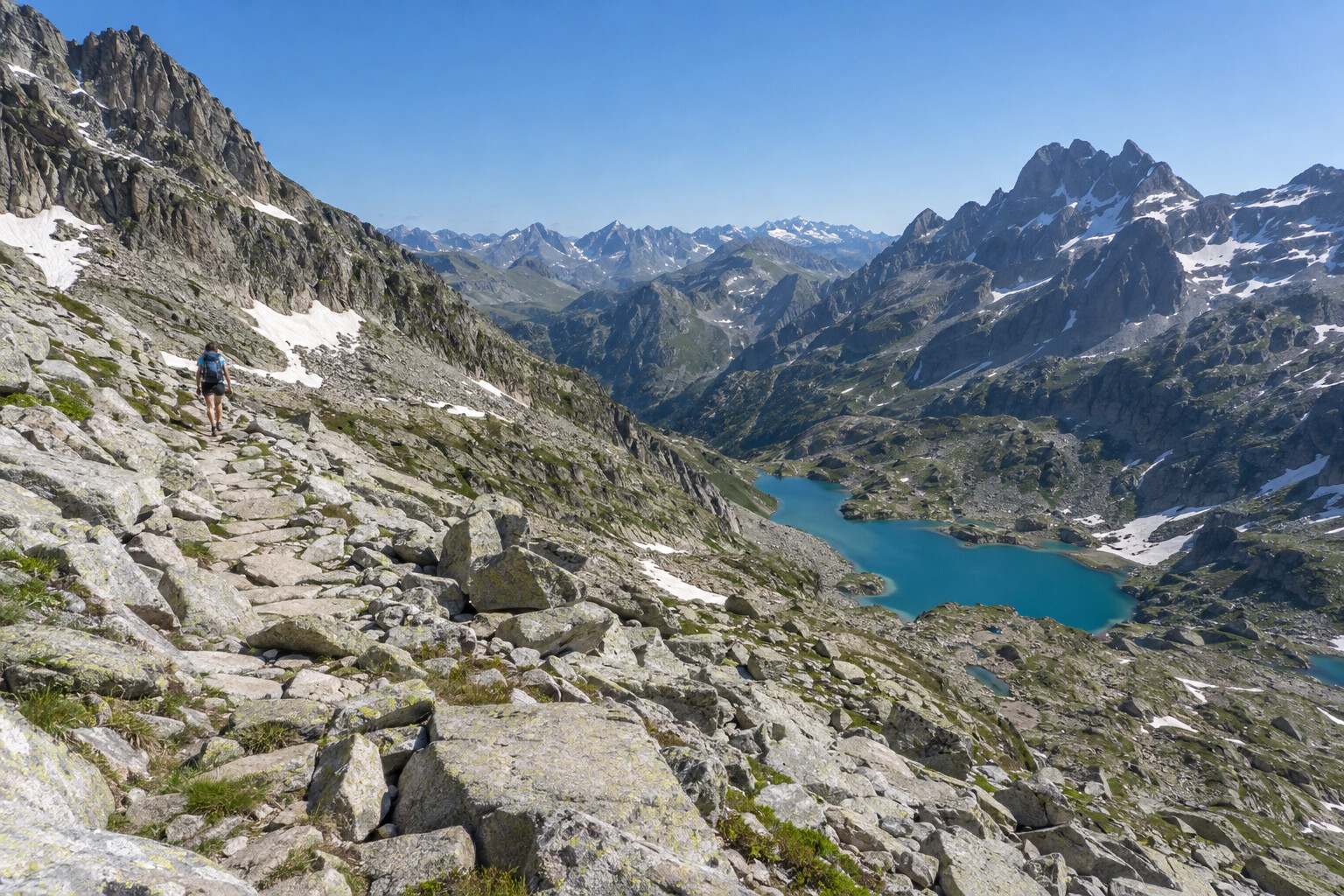Hiker on a granite boulder path between snow patches, above a turquoise high mountain lake on a summer morning