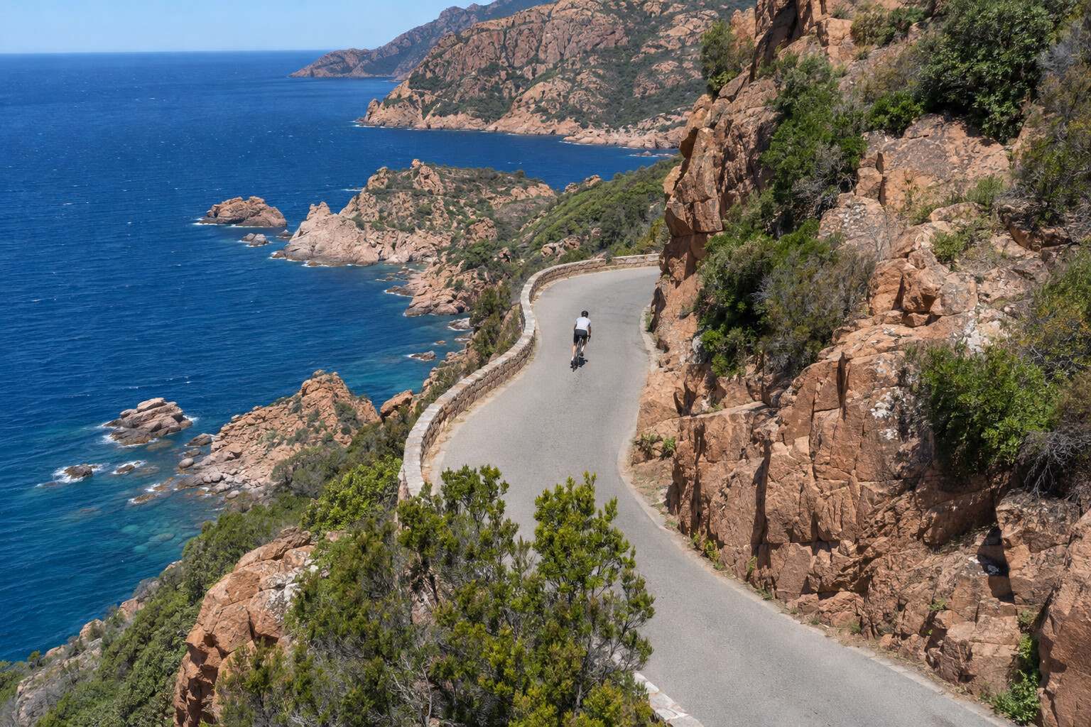 Road cyclist on a coastal road cut into the red rocks of Corsica, high above a deep blue sea